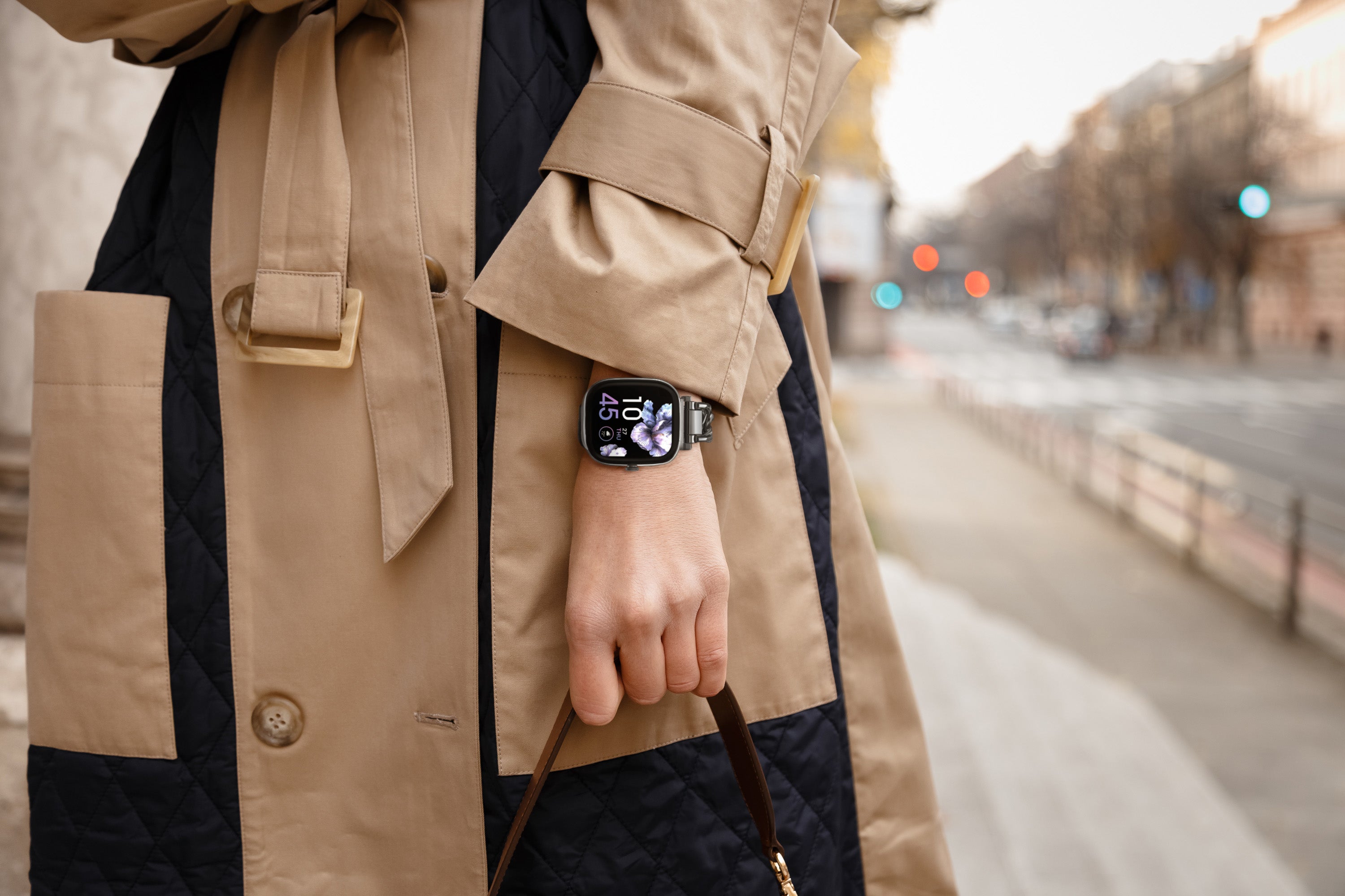 Person wearing a beige trench coat with a blurred city street background wearing smartwatch